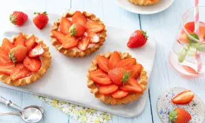 Trois tartelettes fraise aux fraises fraîches, disposées sur un plateau blanc. Une boisson rose avec des fraises et des feuilles de basilic est à droite.