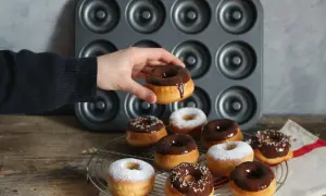 Une main tient un donut glacé au chocolat. Autour, plusieurs donuts, certains saupoudrés de sucre glace, d'autres de glaçage au chocolat et de vermicelles colorés, prêts à être dégustés. Une recette donuts professionnel réussie !