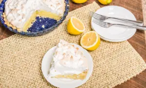 Une part de tarte citron meringuée sur une assiette, à côté d'une tarte entière et de citrons frais. Parfait pour une recette de tarte citron meringuée.