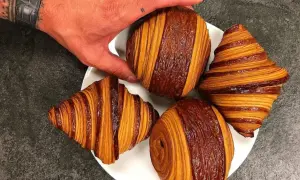 Une main saisit un pain au chocolat Cédric Grolet, aux couches dorées et chocolatées, sur une assiette avec d'autres viennoiseries.