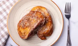 Deux tranches dorées de pain perdu au four, saupoudrées de sucre et de cannelle, sur une assiette avec une serviette à carreaux et une fourchette.