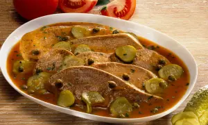 Langue de veau cuisson : fines tranches de langue de veau nappées d'une sauce savoureuse, garnies de cornichons et de câpres. Tomates en arrière-plan.