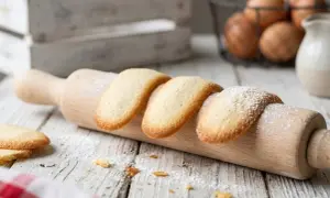 Des biscuits langue de chat dorés, saupoudrés de sucre glace, reposent sur un rouleau à pâtisserie en bois.