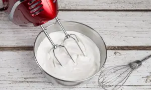 Un batteur rouge prépare une crème chantilly onctueuse dans un bol en inox. Un fouet repose à côté.