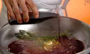 Une main verse du jus de viande dans une poêle avec de l'ail, du romarin et du thym.