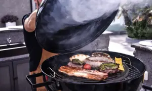 Un homme soulève le couvercle d'un barbecue fumant, révélant des saucisses, des steaks et du bacon. La cuisson lente permet d'attendrir la viande pour un repas savoureux.
