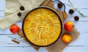 Une tarte aux pommes simple, dorée et appétissante, ornée de fines lamelles de fruits disposées en spirale. Des pommes fraîches, des pommes de pin et une branche de fleurs ajoutent une touche rustique.