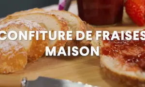 Tranche de pain beurrée et tartinée de confiture de fraise maison, avec une fraise fraîche et un pot de confiture en arrière-plan.