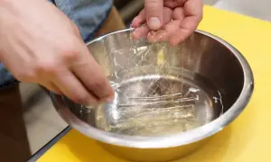 Mains trempant une masse gélatine en feuille dans un bol d'eau. La feuille de gélatine est transparente et a une texture réticulée.