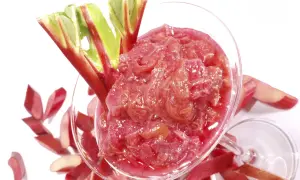 Un verre rempli de compote de rhubarbe, garni de tiges fraîches. Des morceaux de rhubarbe coupés entourent le verre.