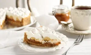 Tranche de tarte bananes et crème fouettée, avec une touche de confiture de lait. Une tasse de thé et une petite jarre complètent ce tableau gourmand.