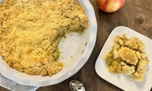 Un délicieux crumble pomme rhubarbe, doré et croustillant, servi dans un plat blanc avec une portion sur une assiette. Une pomme rouge et jaune repose à côté.