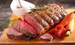 Un rôti de bœuf congelé au four, tranché, avec des carottes, une tomate, de l'ail et du romarin sur une planche en bois.