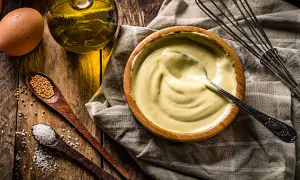 Un bol de mayonnaise maison crémeuse, avec des ingrédients frais : œuf, huile, moutarde et sel. Un fouet à portée de main.