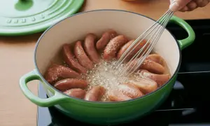 Saucisses de Strasbourg dorées et croustillantes dans une cocotte verte, une main remuant avec un fouet pour une cuisson parfaite.