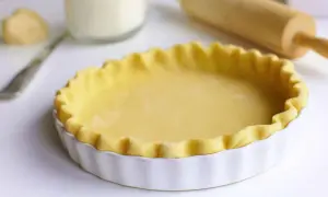 Pâte à tourte prête à être garnie, dans un plat blanc cannelé. Un rouleau à pâtisserie est visible en arrière-plan.