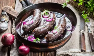 Trois boudins noirs en cuisson dans une poêle, accompagnés d'oignons rouges et de persil. Un plat rustique prêt à être dégusté.