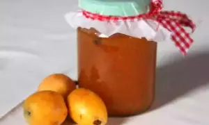 Un pot de confiture de nèfles, décoré d'un ruban à carreaux rouges et blancs, est posé à côté de trois fruits frais.