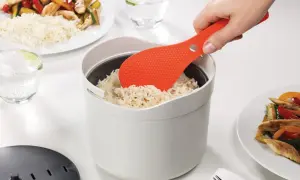 Une spatule orange sert du riz rond dans un cuiseur blanc. Le riz est prêt pour un repas savoureux.