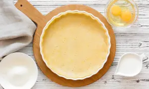 Une pâte à tarte crue est prête dans un plat blanc, entourée d'ingrédients pour différentes pâtes à tarte : œufs, lait, farine.