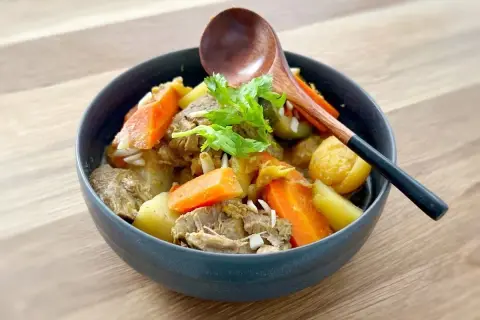 Un délicieux tajine agneau aux carottes et pommes de terre, garni de coriandre fraîche. Une cuillère en bois repose sur le plat.