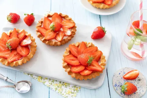 Trois tartelettes fraise aux fraises fraîches, disposées sur un plateau blanc. Une boisson rose avec des fraises et des feuilles de basilic est à droite.
