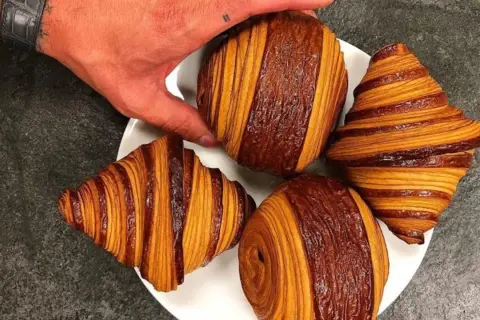 Une main saisit un pain au chocolat Cédric Grolet, aux couches dorées et chocolatées, sur une assiette avec d'autres viennoiseries.