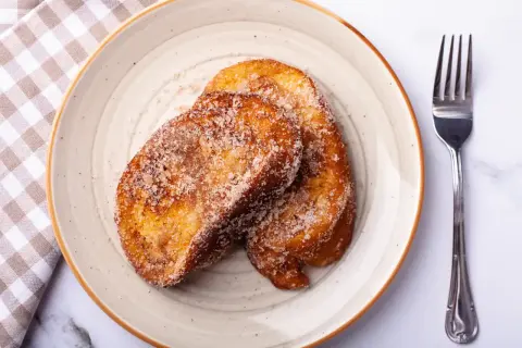Deux tranches dorées de pain perdu au four, saupoudrées de sucre et de cannelle, sur une assiette avec une serviette à carreaux et une fourchette.