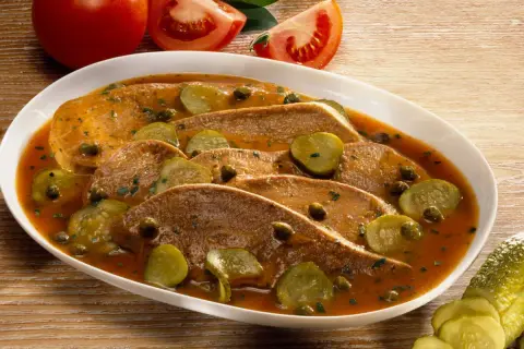 Langue de veau cuisson : fines tranches de langue de veau nappées d'une sauce savoureuse, garnies de cornichons et de câpres. Tomates en arrière-plan.