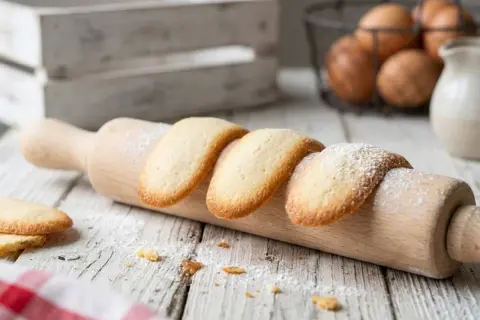 Des biscuits langue de chat dorés, saupoudrés de sucre glace, reposent sur un rouleau à pâtisserie en bois.