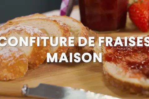 Tranche de pain beurrée et tartinée de confiture de fraise maison, avec une fraise fraîche et un pot de confiture en arrière-plan.