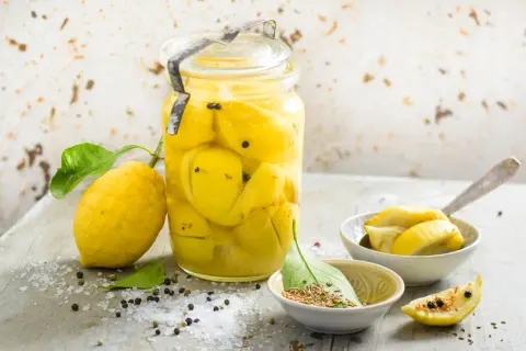 Un bocal de citrons confits au sel, prêts pour une recette citron confit sucre. Des épices et du gros sel parsèment la table.