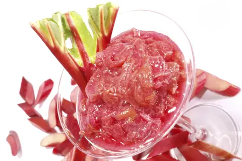 Un verre rempli de compote de rhubarbe, garni de tiges fraîches. Des morceaux de rhubarbe coupés entourent le verre.