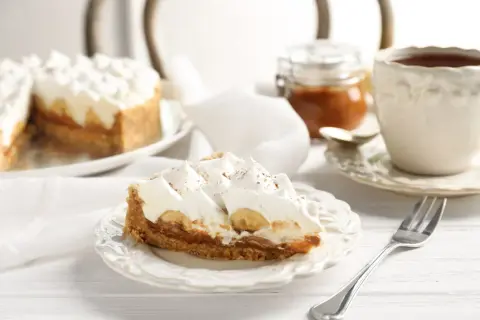 Tranche de tarte bananes et crème fouettée, avec une touche de confiture de lait. Une tasse de thé et une petite jarre complètent ce tableau gourmand.