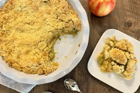 Un délicieux crumble pomme rhubarbe, doré et croustillant, servi dans un plat blanc avec une portion sur une assiette. Une pomme rouge et jaune repose à côté.