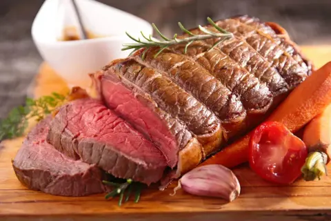 Un rôti de bœuf congelé au four, tranché, avec des carottes, une tomate, de l'ail et du romarin sur une planche en bois.