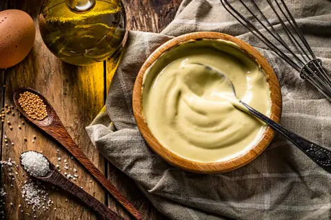 Un bol de mayonnaise maison crémeuse, avec des ingrédients frais : œuf, huile, moutarde et sel. Un fouet à portée de main.
