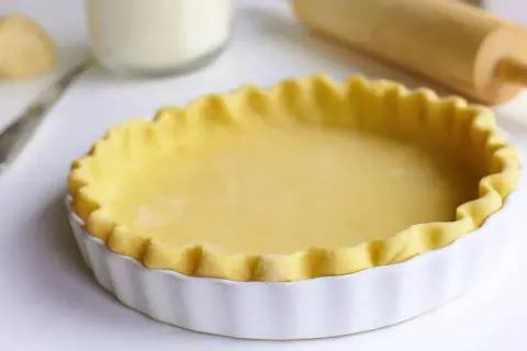 Pâte à tourte prête à être garnie, dans un plat blanc cannelé. Un rouleau à pâtisserie est visible en arrière-plan.