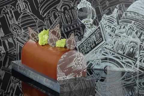 Bûche de Noël chocolatée, décorée de chocolat blanc et de mousse verte, évoquant une recette Pierre Hermé. Le fond présente des croquis architecturaux parisiens.