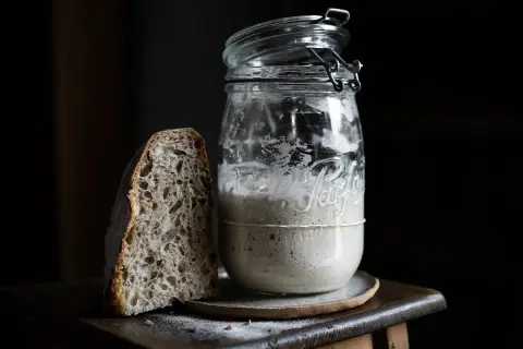 Une tranche de pain au levain maison à côté d'un bocal rempli de levain actif.