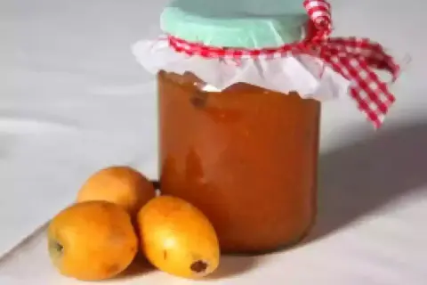 Un pot de confiture de nèfles, décoré d'un ruban à carreaux rouges et blancs, est posé à côté de trois fruits frais.