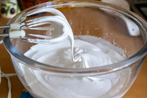 Fouets de batteur électrique en train de battre les blancs en neige dans un bol en verre, créant une texture aérienne parfaite pour une meringue.