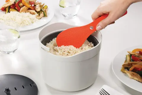 Une spatule orange sert du riz rond dans un cuiseur blanc. Le riz est prêt pour un repas savoureux.