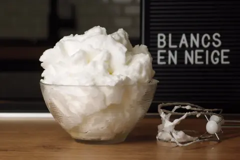 Un bol de blancs en neige aériens, comme un nuage, à côté d'un batteur. Le mot "BLANCS EN NEIGE" est visible sur un panneau.