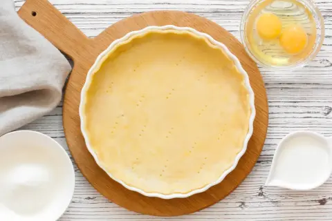 Une pâte à tarte crue est prête dans un plat blanc, entourée d'ingrédients pour différentes pâtes à tarte : œufs, lait, farine.