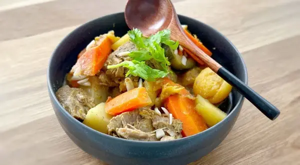 Un délicieux tajine agneau aux carottes et pommes de terre, garni de coriandre fraîche. Une cuillère en bois repose sur le plat.