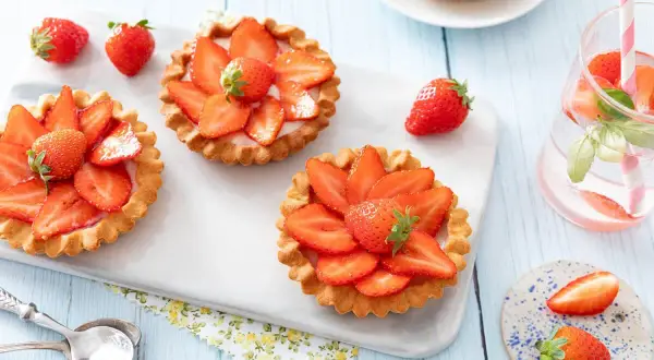 Trois tartelettes fraise aux fraises fraîches, disposées sur un plateau blanc. Une boisson rose avec des fraises et des feuilles de basilic est à droite.