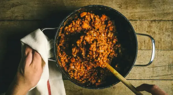 Cuisson lente de lentilles vertes dans une cocotte, avec une cuillère en bois et une main tenant un torchon.