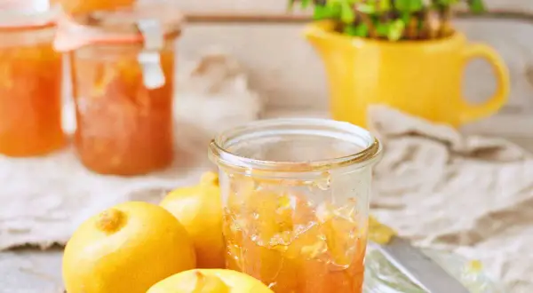 Bocaux de marmelade de citron italienne, avec des citrons frais et une plante verte dans un pichet jaune en arrière-plan.