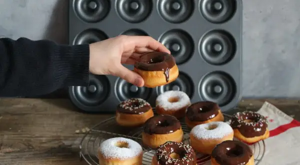 Une main tient un donut glacé au chocolat. Autour, plusieurs donuts, certains saupoudrés de sucre glace, d'autres de glaçage au chocolat et de vermicelles colorés, prêts à être dégustés. Une recette donuts professionnel réussie !